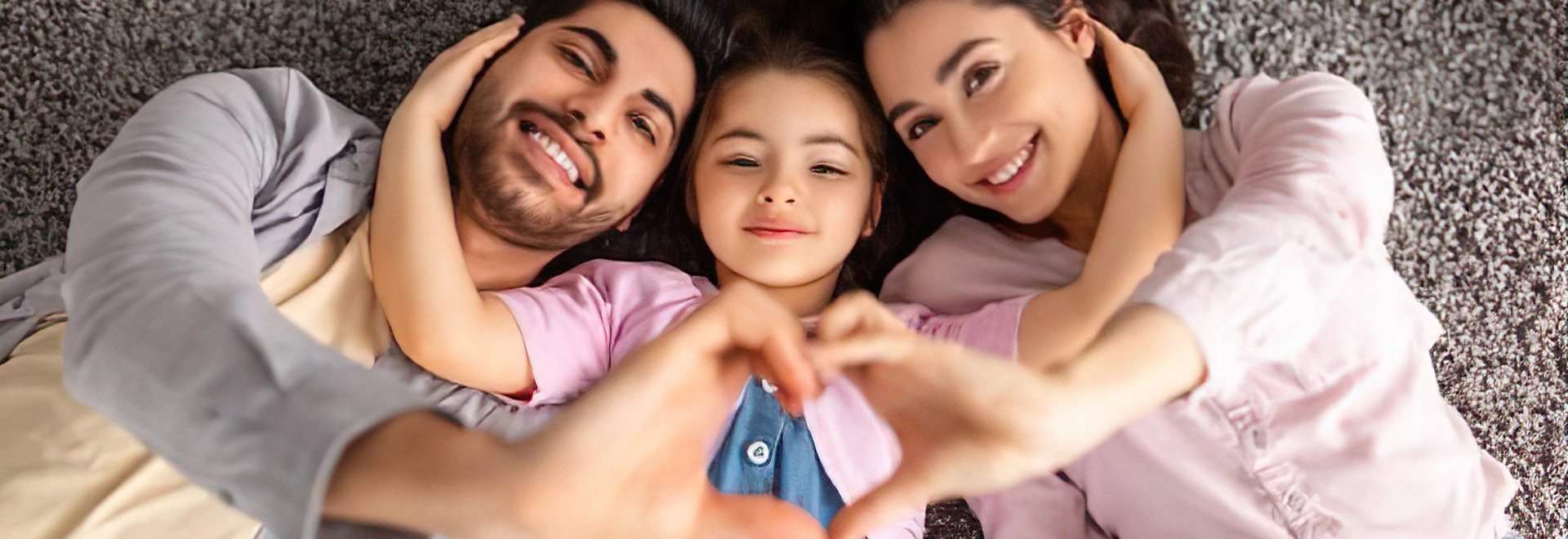 Happy family of three making a heart shape with their hands on a cozy carpet.