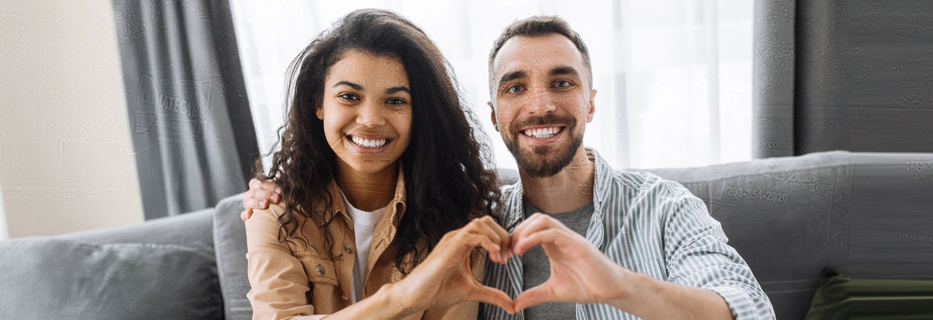 Couple making a heart shape with their hands while smiling on a sofa.