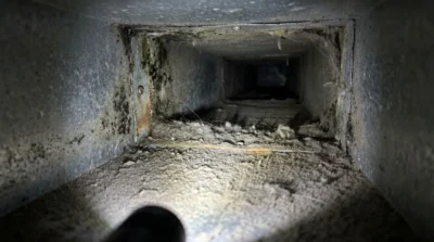 View inside a dusty air duct with cobwebs and debris illuminated by a flashlight.
